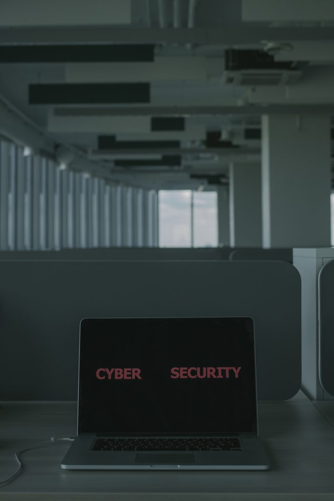 A laptop connected to power displaying a black screen with red text "Cyber Security" located in a cubicle office