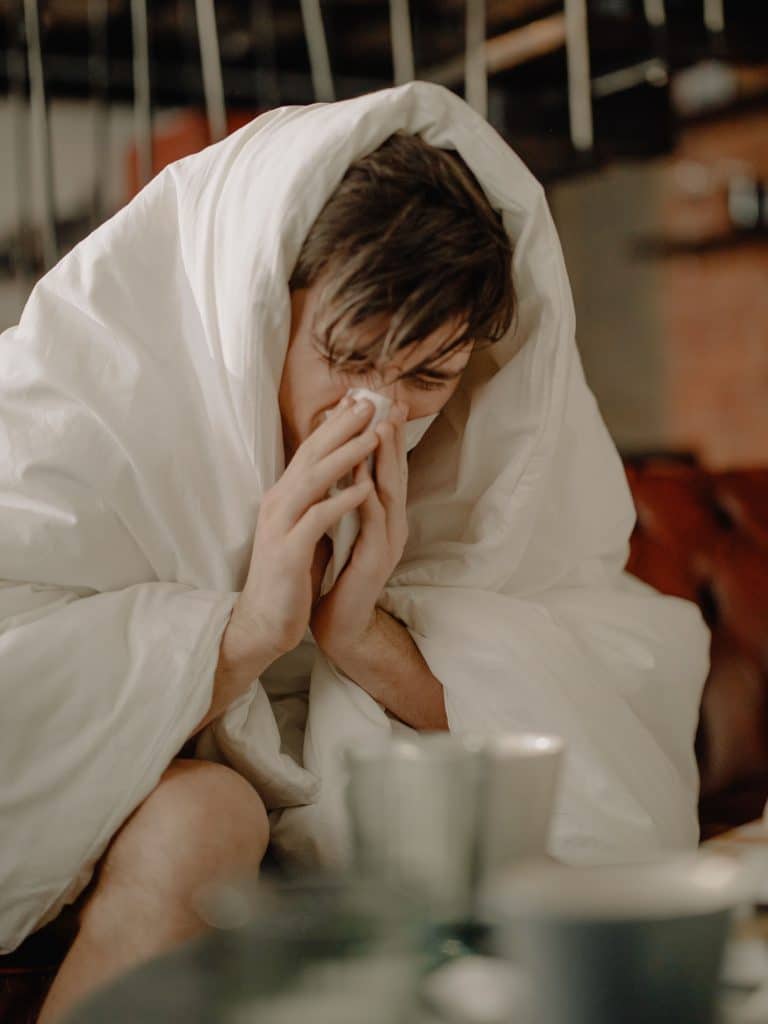 A man blowing his nose while covered in a white blanket sitting in a bed, blurred countertop table with metal dishes